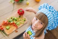 TOP VIEW: Little girl in cook clothes eats a cucumber and looks to the camera
