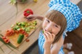 TOP VIEW: Little girl in cook clothes eats a cucumber, smiles and looks to the camera