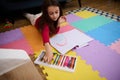 Top view of little girl drawing using pastel crayons while lying on a colorful multicolored puzzle carpet on the floor