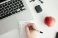 Top view of a man writing on his notebook with a laptop and apple on a white table