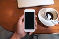 Hand holding white mobile phone with blank black desktop screen with coffee cup and laptop on wooden table in cafe Royalty Free Stock Photo