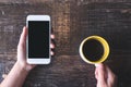 Hand holding white mobile phone with blank black desktop screen and a cup of coffee on wooden table in cafe Royalty Free Stock Photo