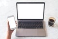 A hand holding white mobile phone with blank screen with laptop with blank white desktop screen and coffee cup on table