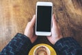 Top view mockup image of hands holding white mobile phone with blank black desktop screen and yellow coffee cup on wooden table Royalty Free Stock Photo