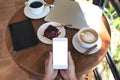 Top view mockup image of hands holding white smartphone with blank screen , tablet , laptop , coffee cup and cake Royalty Free Stock Photo