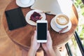 Top view mockup image of hands holding white smartphone with blank screen , tablet , laptop , coffee cup and cake on wooden table Royalty Free Stock Photo