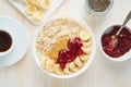 Top view of oatmeal porridge with strawberry jam, peanut butter, banana, coffee on white wooden light background, healthy vegan