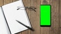 Top view of open notebook with blank pages and cell phone with green screen, glasses on wooden table background for adding text or Royalty Free Stock Photo