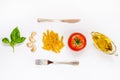Top view of pasta ingredients and cutlery over white background - raw fusilli, fresh basil, garlic cloves, olive oil and ripe toma
