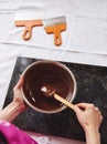 Top view of pastry chef mixing melted chocolate mass with wooden spoon, scrapers lying near marble surface on white tablecloth