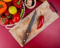 top view of pepper and knife on cutting board with vegetables in basket and garlic crusher on bordo background