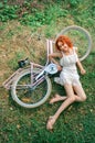 Top view. A pink bike lies on the green grass. A curly red-haired girl lies and laughs nearby. Vertical oriented