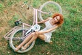 Top view. A pink female bike lies on the green grass. A curly red-haired girl lies and laughs nearby