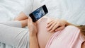 Top view of pregnant woman holding smartphone and looking on ultrasound image of her unborn baby in belly. Concept of Royalty Free Stock Photo