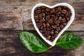 top view of roasted coffee beans in white heart-shaped bowl on wooden background with green leaves of coffea arabica Royalty Free Stock Photo