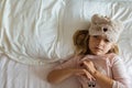 Top view of cute blonde little girl in pajamas and blindfold lying in white bed, awaking early in the morning before going to Royalty Free Stock Photo