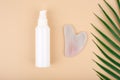 Top view of skin lotion, rose quartz heart shaped guasha massage stone and half of palm leaf on light beige background.