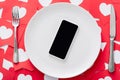 View of smartphone with blank screen on white plate near knife and fork on red paper cut cards with hearts symbols Royalty Free Stock Photo