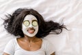 Top-view of smiling African woman making spa procedures with mud facial mask and fresh cucumber slices on eyes, laying Royalty Free Stock Photo