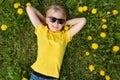 Top view on smiling blond boy wearing sunglasses and yellow T-shirt lying on field of dandelions. Happy childhood Royalty Free Stock Photo