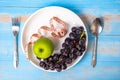 Top view spoon and fork, green apple and black grapes in white dish with pink Measuring tape on blue wood table background.