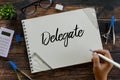 Top view of stationery glasses, calculator,plant and hand holding pen writing Delegate on notebook on wooden background