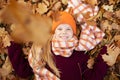 Top view of teenage girl child wearing vinous sweater, orange hat, checkered scarf, lying on yellow fallen maple leaves.