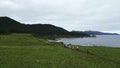 Top view of tourists hiking on green coast on cloudy day. Clip. Hikers walk along coastal path with green mountains and