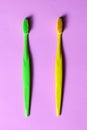 Top view on two colored tooth brushes, green and yellow, laying down on lilac pink background.