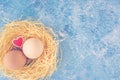 Top view of two fresh chicken eggs in straw nest and decorative wooden heart on blue background. Happy easter greeting Royalty Free Stock Photo