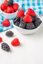 Top view of two white bowls with blackberries and raspberries on blue checkered cloth, on white wooden table, vertically