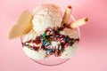 Top view of vanilla flavor ice cream in glass bowl with chocolate sauce, strewed sprinkles and waffle cookies on pink background