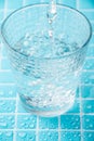 Top view of water jet falling into a glass cup, with selective focus, on wet light blue mosic, in vertical, Royalty Free Stock Photo