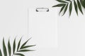 Top view of a white clipboard mockup with palm leaf decoration on a white table