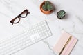 Top view of white keyboard, glasses, pink notebook and cactus on marble background in horizontal flatlay with negative space. Royalty Free Stock Photo