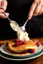 Top view of woman`s hands with spoons and mascarpone cheese on pancakes with cherries