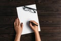 Top view of women`s hands, ready to write something on an empty piece of paper lying on a wooden table. Write a letter on a white