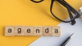 Top view the word of the agenda written on wooden cubes on the desktop with glasses, pen, paper Notepad on a yellow background, Royalty Free Stock Photo