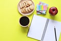 Top view of working desk with blank notebook with pencil, cookies, apple, coffee cup and colorful note pad on yellow background