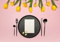 Top view of yellow tulips, daffodils, easter eggs on pink background. Table setting, black plate and cutlery, white card. Royalty Free Stock Photo