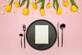 Top view of yellow tulips, daffodils, easter eggs on pink background. Table setting, black plate and cutlery, white card. Royalty Free Stock Photo