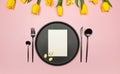 Top view of yellow tulips, daffodils, easter eggs on pink background. Table setting, black plate and cutlery, white card. Royalty Free Stock Photo