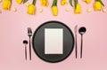 Top view of yellow tulips, daffodils, easter eggs on pink background. Table setting, black plate and cutlery, white card. Royalty Free Stock Photo