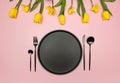 Top view of yellow tulips and daffodils on pink background. Table setting, black plate and cutlery. Royalty Free Stock Photo