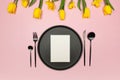 Top view of yellow tulips and daffodils on pink background. Table setting, black plate and cutlery, white card. Royalty Free Stock Photo