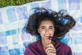 Top view of young brunette woman smiling with lollipop in hand wearing plaid shirt lying on the grass in the park enjoying