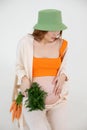 Young stunning pregnant woman with long dark hair wear green bucket hat, sitting, holding belly and bunch of carrots. Royalty Free Stock Photo