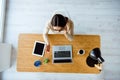 Top-view of young successful woman in glasses sitting at table with laptop, cup of coffee and tablet in office while Royalty Free Stock Photo