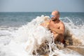 Topless vacationist in sunglasses with drink in hand, screwed eyes while sea wave covering tourist sitting on shore.