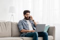 Always in touch. Young arab freelancer man talking on smartphone and using laptop working remotely from home Royalty Free Stock Photo